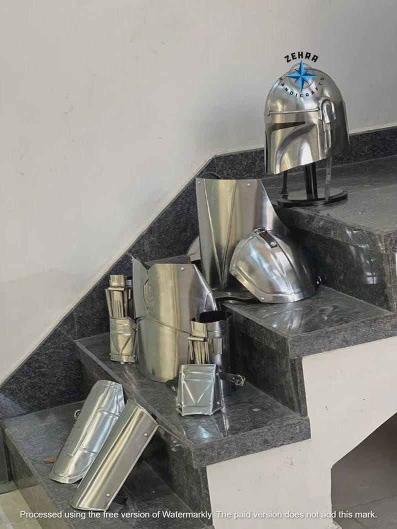 May include: A collection of shiny, silver-colored Mandalorian armor pieces, including a helmet, chest plate, and arm guards. The helmet is displayed on a black stand. The armor is arranged on a set of gray steps.