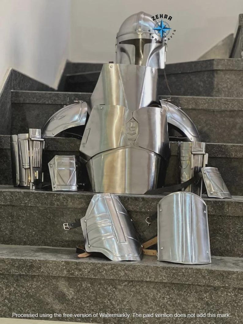 May include: A complete set of silver-colored Mandalorian armor, including a helmet, chest plate, shoulder pads, and gauntlets. The armor is displayed on a set of gray steps, showcasing its intricate design and metallic finish. The image includes the text "ZEHRA HAND CRAFTS".