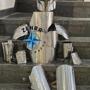 May include: A complete set of metallic silver Mandalorian armor, including a helmet, chest plate, shoulder pads, and various other protective pieces. The armor is displayed on a set of gray steps. The text "ZEHRA HANDICRAFTS" is visible.
