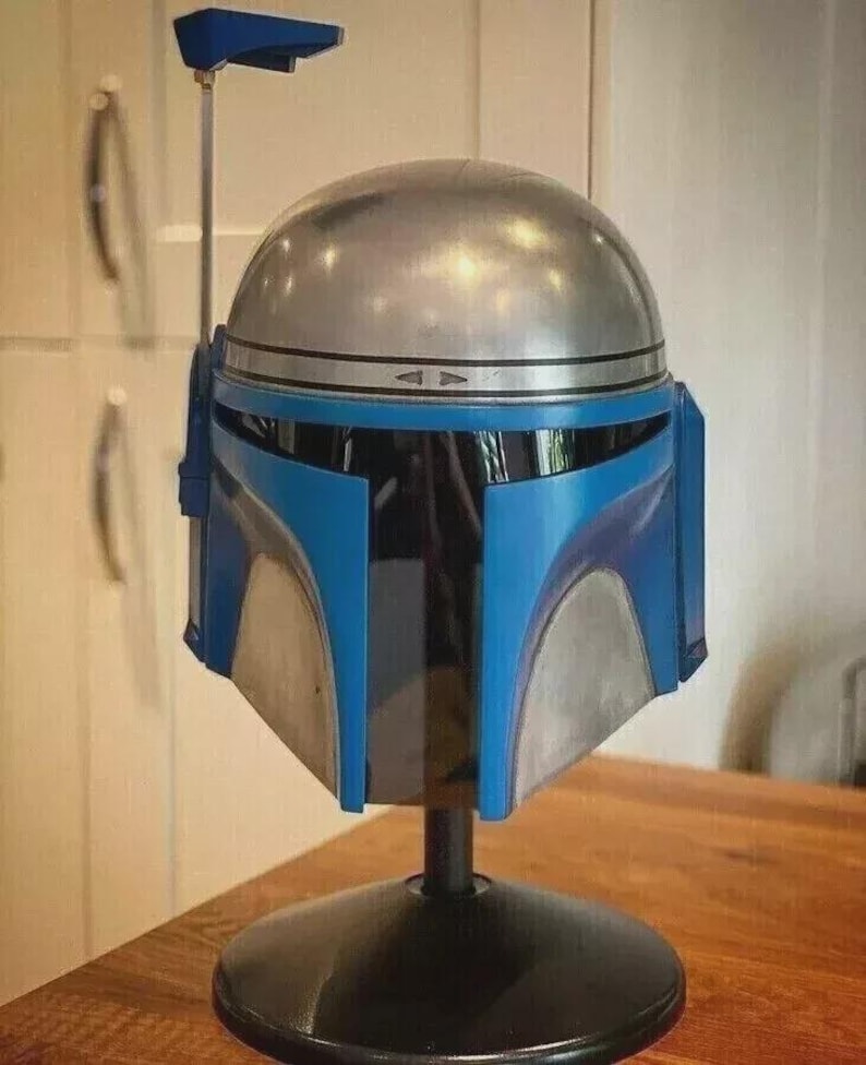 May include: A metallic silver and blue helmet, possibly a prop or collectible, is displayed on a black stand. The helmet features a rounded top, blue accents, and a visor. The design suggests a science fiction or fantasy theme.