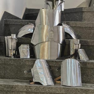May include: A complete set of shiny, silver-colored Mandalorian armor, including a helmet, chest plate, shoulder pads, and gauntlets. The armor is displayed on a set of stairs, showcasing its intricate design and metallic finish.