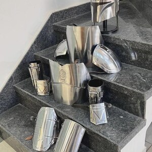 May include: A complete set of polished metal armor, including a helmet, chest plate, shoulder pads, and arm guards, displayed on a staircase. The helmet is on a black stand. The armor is silver and reflects the light.