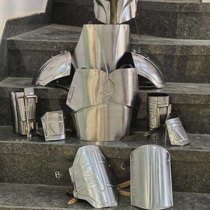 May include: A complete set of polished silver armor, including a helmet, chest plate, shoulder pads, and various other protective pieces. The armor is displayed on a set of gray steps, showcasing its intricate design and metallic sheen.