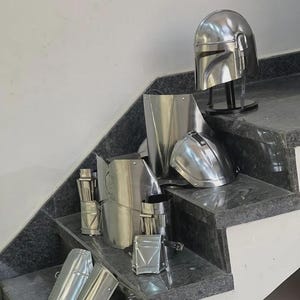 May include: A collection of shiny, silver-colored Mandalorian armor pieces, including a helmet, chest plate, and arm guards, arranged on a set of granite stairs. The armor appears to be made of metal and is displayed in a well-lit environment.