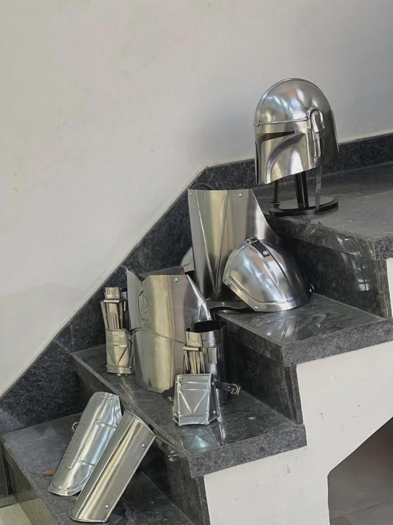 May include: A collection of shiny, silver-colored Mandalorian armor pieces, including a helmet, chest plate, and arm guards, arranged on a set of granite stairs. The armor appears to be made of metal and is displayed in a well-lit environment.