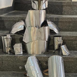 May include: A complete set of polished metal armor, including a helmet, chest plate, shoulder guards, arm and leg pieces. The armor is displayed on a set of gray steps, showcasing its intricate design and reflective surface.