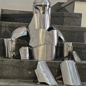 May include: A complete set of silver-colored Mandalorian armor, including a helmet, chest plate, shoulder pads, and arm guards. The armor is displayed on a set of stairs, showcasing its detailed design and metallic finish. This set is perfect for cosplay or display.