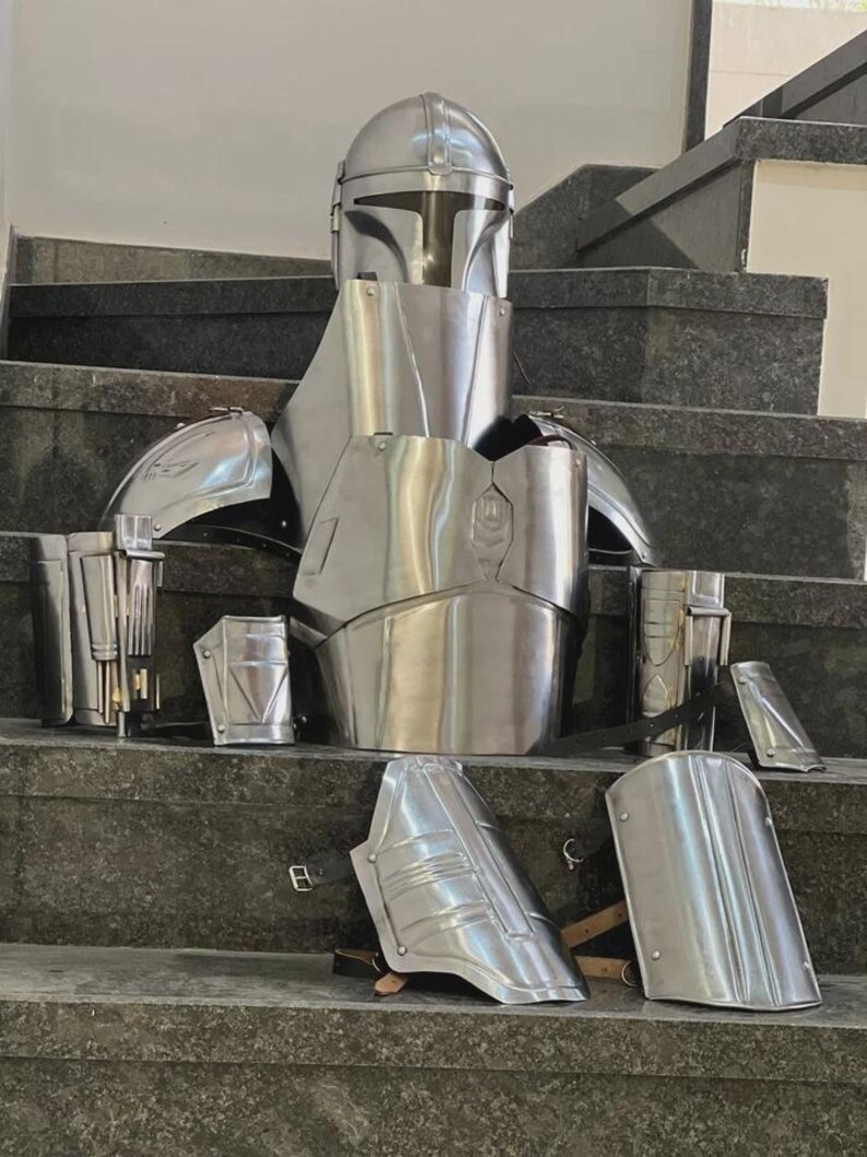 May include: A complete set of silver-colored Mandalorian armor, including a helmet, chest plate, shoulder pads, and arm guards. The armor is displayed on a set of stairs, showcasing its detailed design and metallic finish. This set is perfect for cosplay or display.