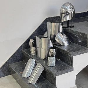 May include: A collection of shiny, silver-colored armor pieces, including a helmet, chest plate, shoulder pads, and arm guards. The armor is displayed on a set of granite stairs, showcasing a futuristic or sci-fi theme.