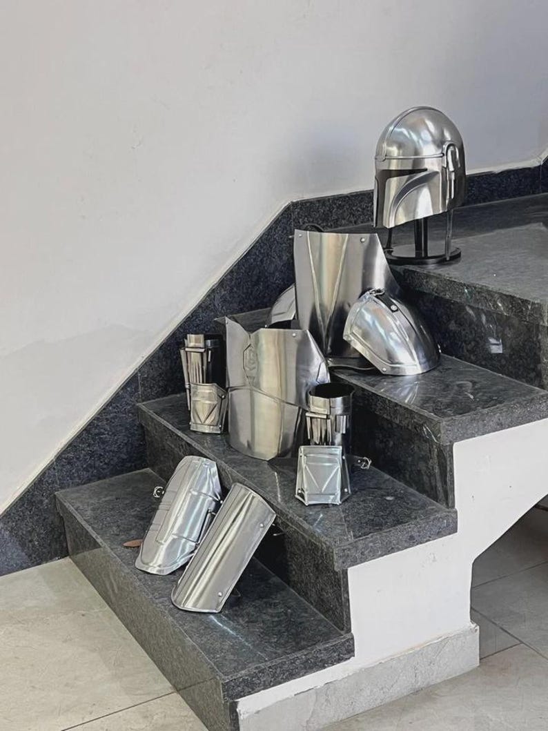 May include: A collection of shiny, silver-colored armor pieces, including a helmet, chest plate, shoulder pads, and arm guards. The armor is displayed on a set of granite stairs, showcasing a futuristic or sci-fi theme.