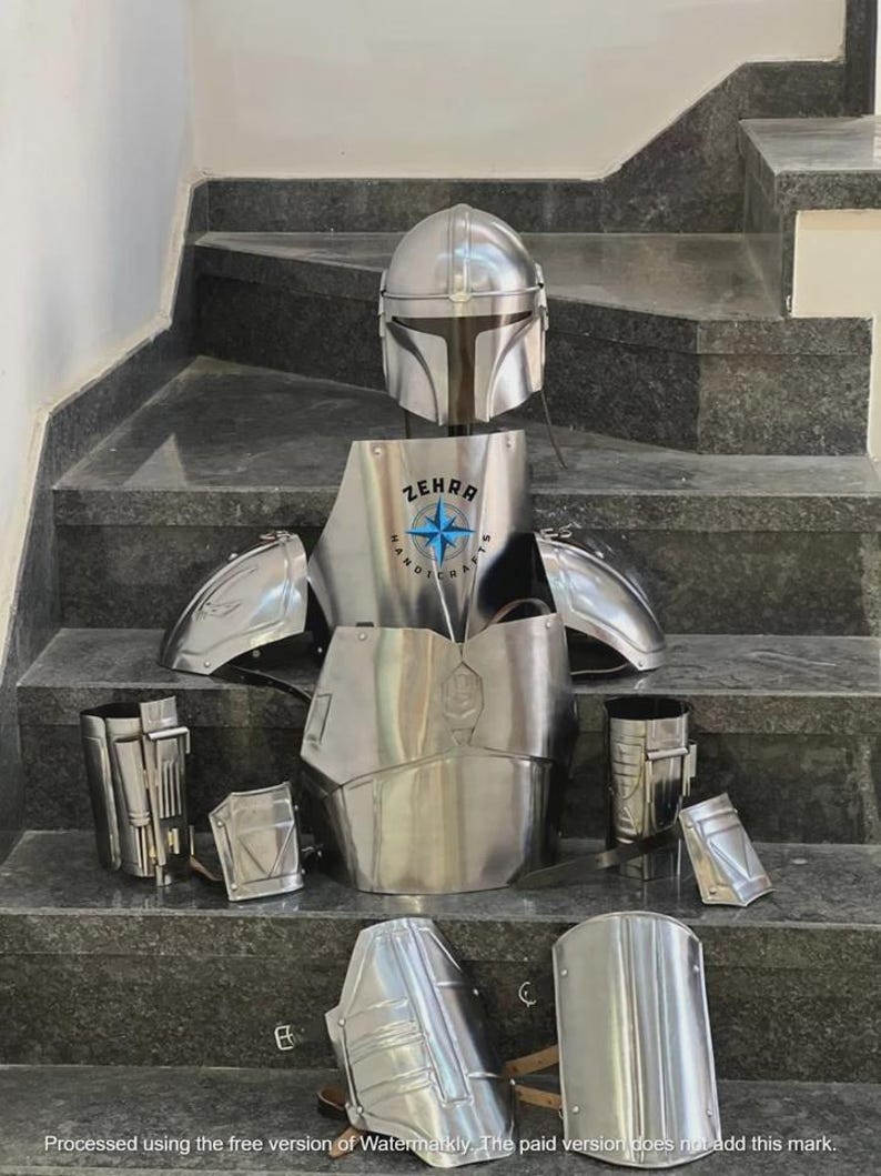 May include: A complete set of polished metal armor, including a helmet, chest plate, shoulder pads, and leg and arm guards. The chest plate features the text "ZEHRA HANDICRAFTS" with a star emblem. The armor is displayed on a set of stairs.