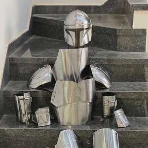 May include: A complete set of polished silver Mandalorian armor, including a helmet, chest plate, shoulder pads, and various leg and arm pieces. The armor is displayed on a set of gray stairs, showcasing its intricate design and reflective surface.