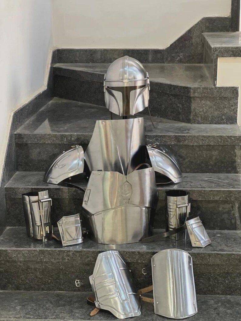 May include: A complete set of polished silver Mandalorian armor, including a helmet, chest plate, shoulder pads, and various leg and arm pieces. The armor is displayed on a set of gray stairs, showcasing its intricate design and reflective surface.