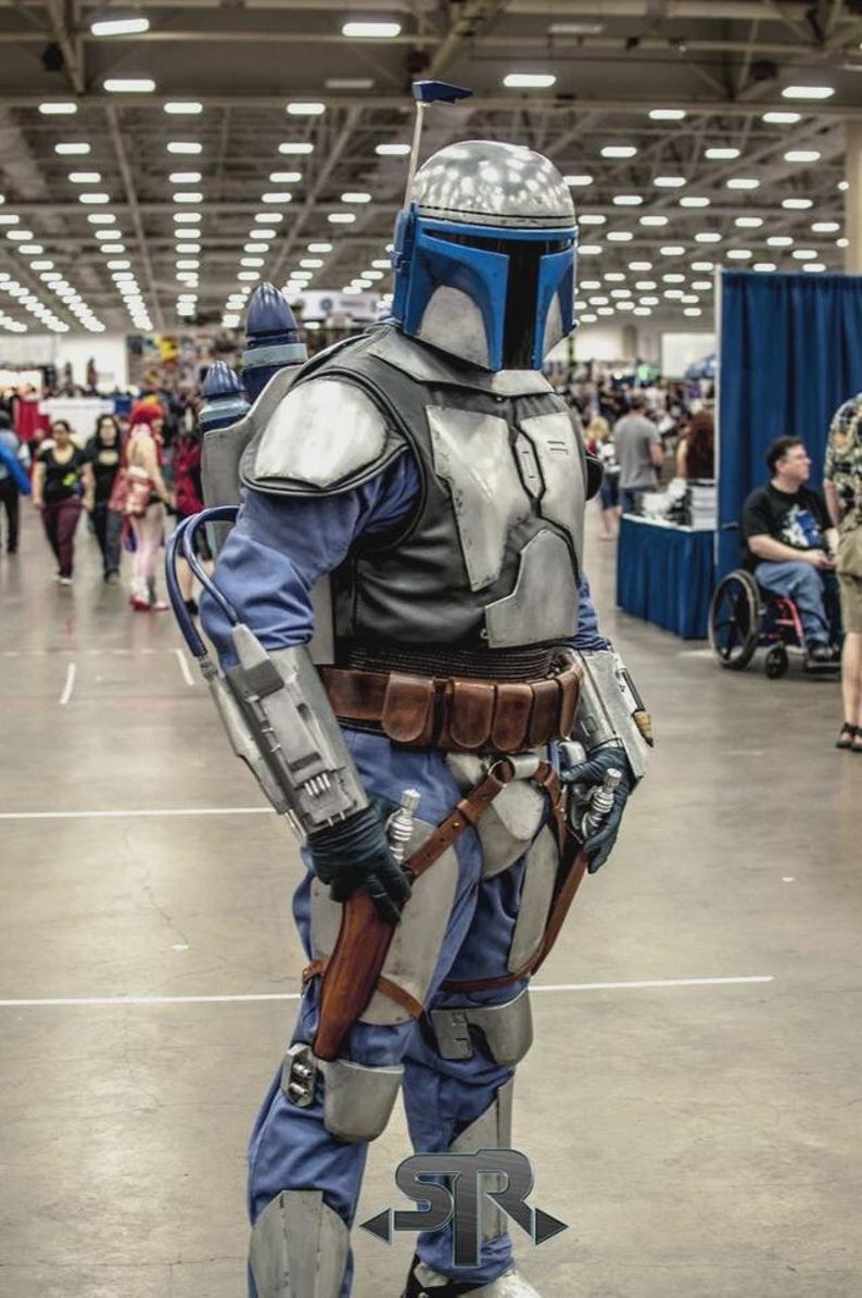May include: A person in a detailed Mandalorian costume, featuring a silver and blue helmet, armor, and a jetpack. The costume includes a blue jumpsuit, brown belt, and various weapon accessories. The setting appears to be a convention hall.