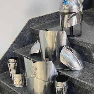 May include: A set of shiny, silver-colored Mandalorian armor pieces, including a helmet, chest plate, shoulder pads, and arm guards. The helmet is displayed on a black stand. The armor is arranged on a set of gray stairs. The brand name "ZEHRA HANDICRAFTS" is visible.
