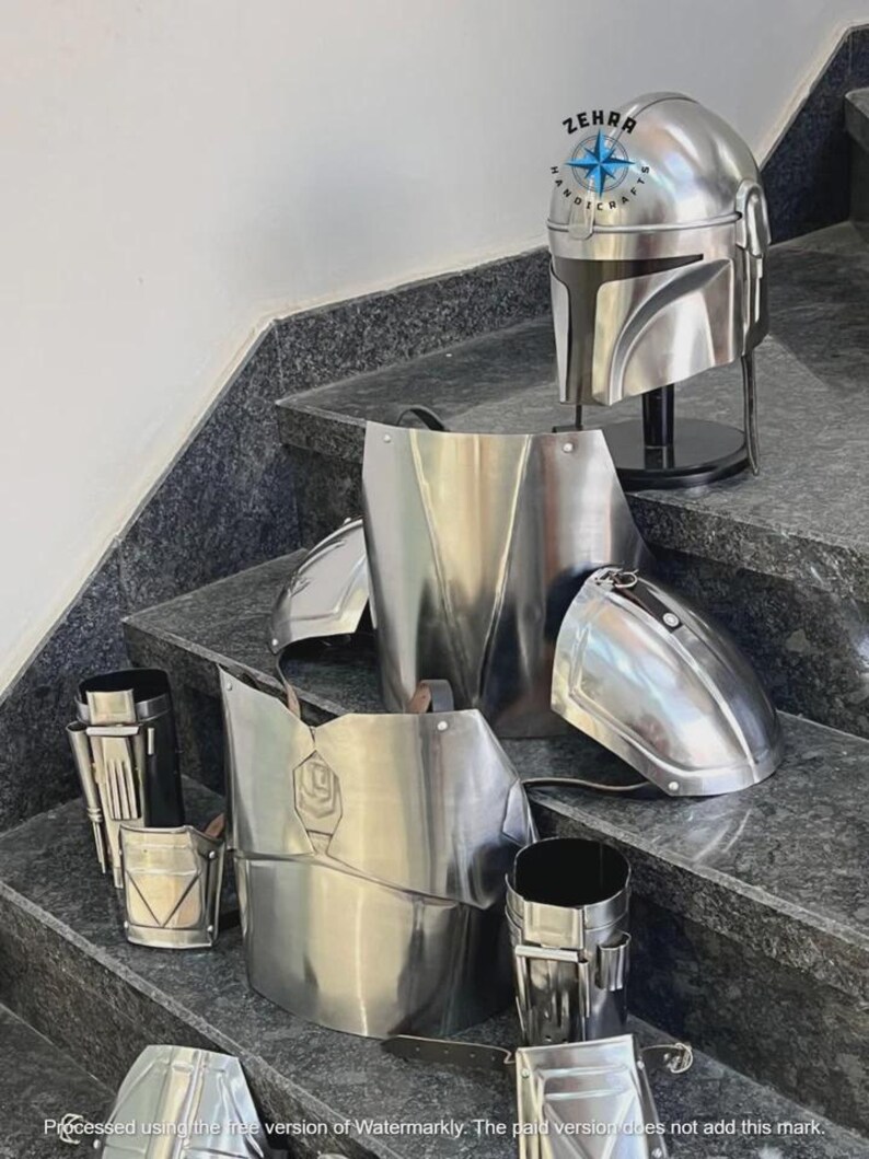 May include: A set of shiny, silver-colored Mandalorian armor pieces, including a helmet, chest plate, shoulder pads, and arm guards. The helmet is displayed on a black stand. The armor is arranged on a set of gray stairs. The brand name "ZEHRA HANDICRAFTS" is visible.