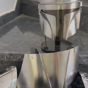 May include: A silver-colored Mandalorian helmet and armor set. The helmet has a T-shaped visor and a rounded top. The armor includes a chest plate and other pieces, all with a metallic finish. The set is displayed on a black stand.