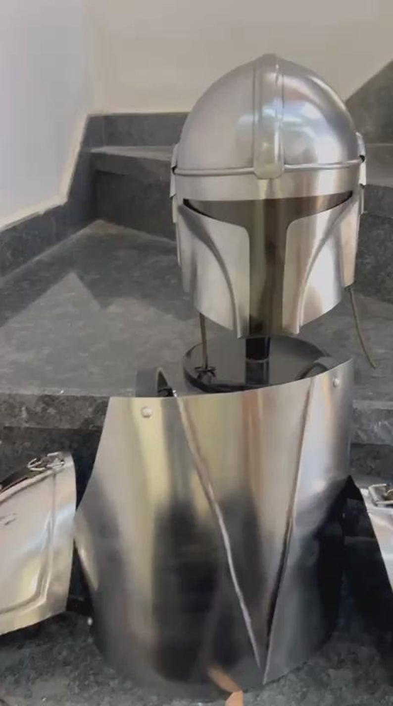 May include: A silver-colored Mandalorian helmet and armor set. The helmet has a T-shaped visor and a rounded top. The armor includes a chest plate and other pieces, all with a metallic finish. The set is displayed on a black stand.