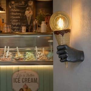 May include: A wall-mounted light fixture featuring a dark-colored hand sculpture holding an ice cream cone. The cone is topped with a large, round, glowing Edison-style light bulb that emits a warm, golden light, creating a unique illumination.