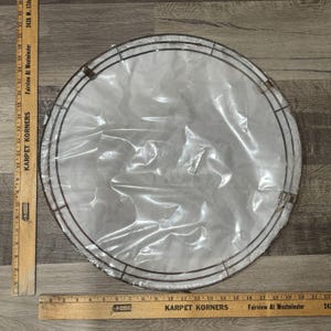 Puede incluir: Un marco de corona circular de metal con acabado oscuro, envuelto en plástico transparente. El marco consta de múltiples anillos concéntricos y está colocado sobre una superficie de madera. Se ven dos reglas de madera.