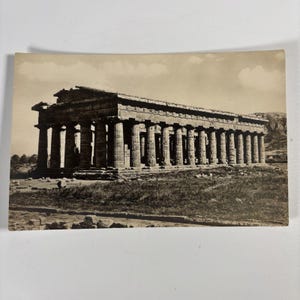 May include: A sepia-toned vintage postcard featuring a wide-angle view of an ancient Greek temple with numerous fluted columns, a triangular pediment, and a stone entablature, set against a landscape with a distant hill under a cloudy sky.
