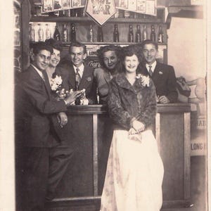 May include: Vintage black and white photograph of a group of people at a bar. The bar has a sign that says "OWL BAR". The people are dressed in formal attire, and the woman in the center is wearing a fur coat and long dress.