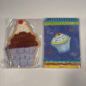May include: Two spiral-bound notebooks in clear plastic packaging. One is shaped like a chocolate cupcake with white frosting and a red cherry. The other is a rectangular blue notebook featuring a cupcake illustration and the text Enjoy Life.