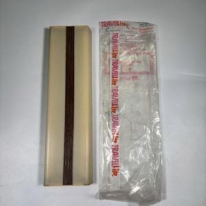 Op de afbeelding: Een rechthoekige lamp met een crèmekleurige doorschijnende kap en een donkerbruine strook met houtnerf in het midden. Daarnaast ligt een doorzichtige plastic verpakking met de rode tekst TRAVELite voor een 12-volt lamp voor recreatievoertuigen.