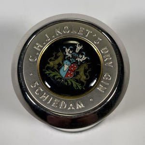 May include: A silver bottle stopper with the text "C.H.J. NOLET'S DRY GIN SCHIEDAM" around a central emblem. The emblem shows a coat of arms with a shield, two lions, and decorative elements.