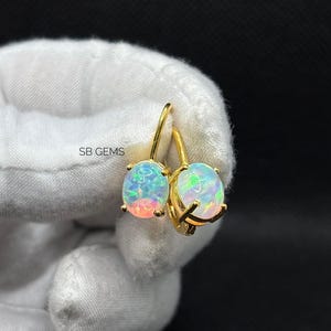 Peut inclure: Une paire de boucles d'oreilles dorées avec des pierres d'opale iridescentes. Les opales présentent un mélange de couleurs, dont le bleu, le vert et le rose. Les boucles d'oreilles sont tenues par une main gantée sur fond sombre. Le texte "SB GEMS" est visible.