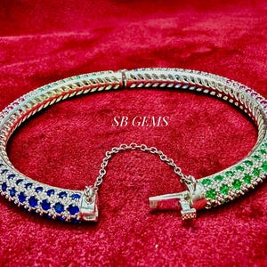 May include: Silver-toned bangle bracelet with a row of blue, green, and red gemstones. The bracelet is open and has a chain attached to the clasp. The text "SB GEMS" is visible on the bracelet.