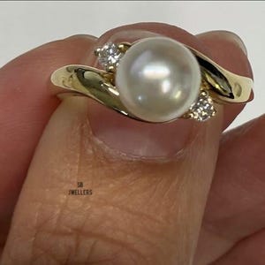 Puede incluir: Un anillo de oro con una perla blanca grande y dos piedras de acento más pequeñas. La banda del anillo tiene un diseño fluido. El anillo está sostenido por los dedos de una persona. La perla es el foco principal del anillo.