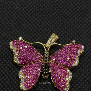 May include: A gold-toned butterfly pendant with pink and white gemstones. The butterfly's wings are filled with pink stones, outlined with white stones. The body is black, and the pendant has the text "SB GEMS".