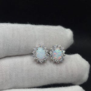 Puede incluir: Un par de pendientes de ópalo con un halo de pequeñas piedras preciosas transparentes. Los ópalos tienen una base blanca lechosa con destellos de color. Los pendientes están engarzados en un metal color plata. El texto "SB GEME" es visible.