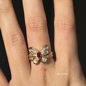 Op de afbeelding: Een gouden ring met een vlinderontwerp. De ring heeft een centrale robijn en is versierd met kleinere diamanten. De ring zit om een vinger. De tekst "SB GEMS" is zichtbaar.