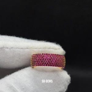 Op de afbeelding: Een goudkleurige ring bezet met talrijke kleine, levendige roze edelstenen. De ring wordt getoond tegen een witte stoffen achtergrond, met de tekst "SB GEMS" zichtbaar. Het ontwerp van de ring heeft een doorlopende band van de roze stenen.