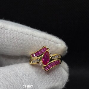 Pode incluir: Um anel de ouro com uma pedra central de rubi em forma de marquesa, ladeado por rubis em corte baguete e pequenos diamantes. O anel tem um design assimétrico único. O texto "SB GEMS" é visível.