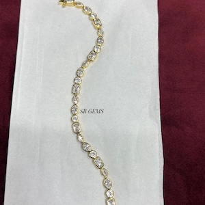 May include: A gold bracelet featuring clear, rectangular and oval-cut gemstones. The bracelet is displayed on a white surface with the text "SB GEMS". The background is a dark red colour.