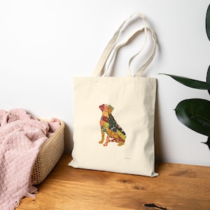 May include: A cream-coloured tote bag with a patchwork dog design. The dog is sitting, made up of floral and geometric patterns in red, yellow, green, and blue. The bag has two handles, perfect for carrying items.