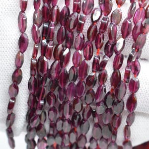 May include: Close-up of multiple strands of oval-cut, deep burgundy garnet beads. The beads are strung on red thread. The polished gemstones have a translucent quality, reflecting light. Suitable for jewellery making.