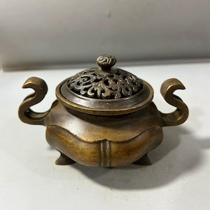 May include: A bronze-colored incense burner with a decorative, pierced lid. The burner has curved handles on either side and rests on three small feet. The lid is topped with a small, ornate knob. The overall design is traditional and elegant.