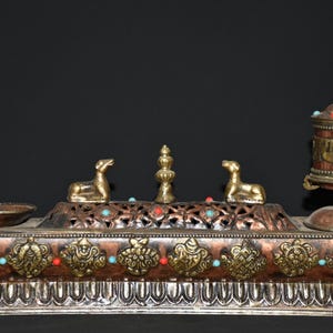 May include: An ornate, multi-piece metal religious artifact. The piece features a rectangular base with decorative carvings, gold accents, and turquoise and red stone inlays. Two golden animal figures and a prayer wheel are also present.