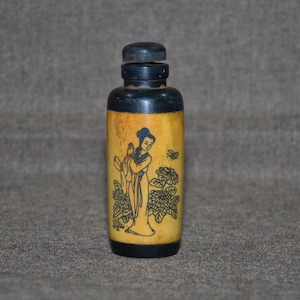 Op de afbeelding: Een kleine, antieke fles met een zwarte dop en bodem. De fles heeft een lichtgele kleur en een zwarte illustratie van een vrouw, bloemen en een bij. De fles heeft een klassieke, vintage uitstraling.