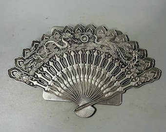 May include: A silver-colored fan-shaped decorative object. The fan features intricate relief carvings of a dragon and a phoenix, with cloud and floral motifs. The fan has a rectangular base and a detailed, openwork design.