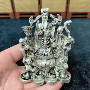 May include: A detailed silver figurine depicting three figures with long beards and ornate clothing. The central figure is seated, flanked by two standing figures, and surrounded by smaller figures and vessels. The figurine is held in a hand.