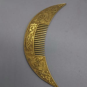 May include: A gold-colored, crescent-shaped comb with intricate dragon and floral designs. The comb's handle features detailed carvings, and the teeth are evenly spaced. The overall design suggests a vintage or antique aesthetic.
