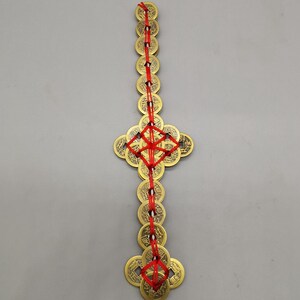 May include: A decorative hanging ornament featuring multiple brass-colored coins strung together with red cord. The coins are arranged in a vertical line and a cross-like shape, creating a symmetrical design.