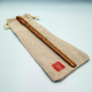 May include: A wooden chopstick with a textured, brown grain pattern rests on a beige linen pouch. The pouch has a drawstring closure and a red square stamp. The chopstick is a dining utensil.