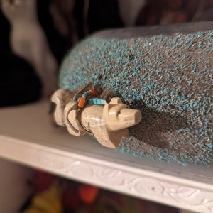 May include: A small white carved animal figurine with turquoise and orange beads around its neck. The figurine is sitting on a turquoise and gray textured surface.