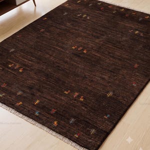 May include: A rectangular dark brown area rug featuring a subtle, textured surface with small, scattered multi-colored geometric motifs and thin, light-colored fringe detailing along the two shorter edges of the woven floor covering.